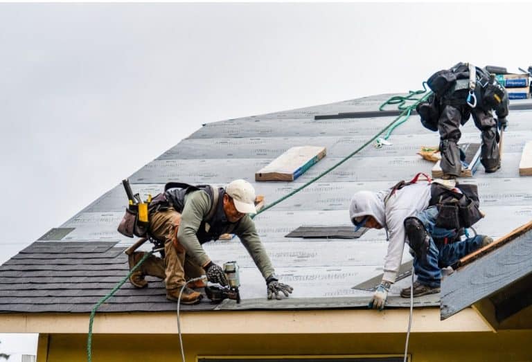 Roofing professionals repair a residential roof by installing new shingles. Workers use safety harnesses and tools while replacing damaged roofing materials.